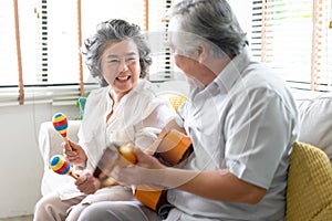 Portrait of smiling senior couple playing guitar singing and her wife holding maracas dancing and sitting on sofa at home.