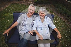 Portrait of smiling senior couple sitting with laptop on couch in backyard