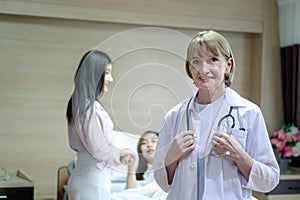 Portrait of smiling senior female doctor in workwear with stethoscope standing in recovery room at clinic hospital with patient