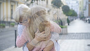 Portrait of smiling senior grandmother in eyeglasses hugging blond little granddaughter outdoors. Happy Caucasian woman
