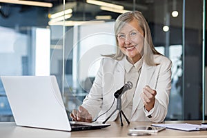 Portrait of a smiling senior gray-haired business woman sitting in the office at a desk in front of a microphone and a