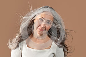Portrait of smiling senior woman posing with long and healthy grey blowing flying hair on brown studio background