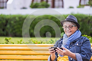 Portrait of smiling senior woman sitting on a yellow bench in public park, using cellphone. Old people with hat modern social and