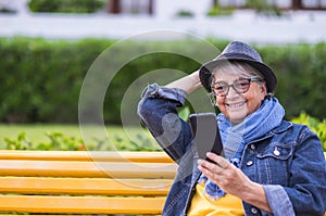 Portrait of smiling senior woman sitting on a yellow bench in public park, using cellphone. Old people with hat modern social and