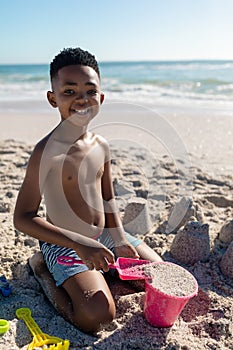 Portrait of smiling shirtless african american boy playing with sand pail and shovel at beach