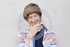 Portrait of smiling shy boy with trendy hairdo wearing casual shirt looking down holding hand on chin having dimple on his cheek.