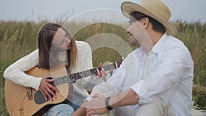 Portrait of smiling skilled young woman playing guitar as blurred man singing sitting on picnic in autumn field