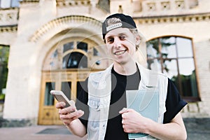 Portrait of a smiling student using a mobile phone and holds a notebook on the background of the university building. photo
