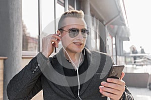 Portrait of a smiling stylish guy of Caucasian appearance of a businessman in sunglasses, jacket and shirt, adjusts his headphones