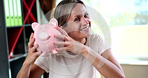 Portrait of smiling successful young woman cracking a piggy bank in hands in office.