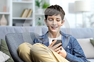 Portrait of a smiling teenage boy child sitting on the couch at home and using the phone. Chatting, sits in social
