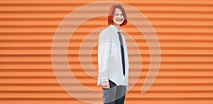 Portrait of smiling teenage girl with painted red dyed hair in white school shirt and necktie standing near orange wall background