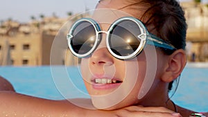 Portrait of a smiling teenage girl, wearing funny, round sunglasses, relaxing in outdoor pool of a posh hotel. sunrise