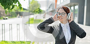 Portrait of a smiling woman in a business suit listening to music in headphones outdoors. Female office employee holds