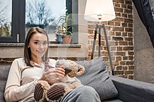 portrait of smiling woman with cup of drink and teddy bear in hands looking at camera while resting on sofa