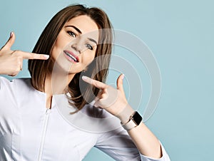 Portrait of smiling woman dentist or orthodontist in white medical uniform gown pointing at lower jaw with both hands