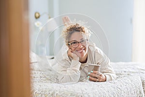 Portrait of smiling woman in eyeglasses and curly hair lying on comfortable bed with coffee cup at home. Happy woman having