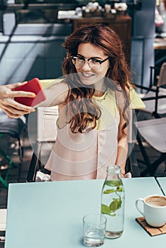 portrait of smiling woman in eyeglasses taking selfie on smartphone