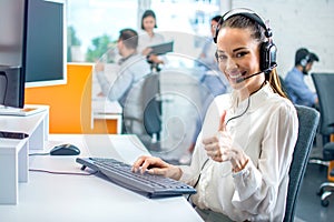 Portrait of smiling woman with headset showing approve sign with thumb up to camera at call center office