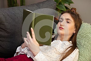 Portrait of smiling woman holding book and lying on couch. Teenager girl home - student read book laying on sofa