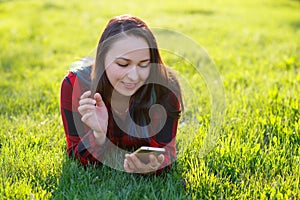 Portrait of a smiling woman lying on green grass and using smartphone outdoors