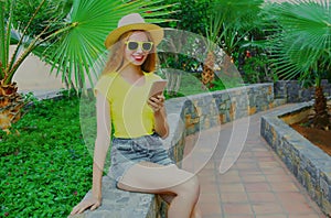 Portrait smiling woman with phone sitting in a park wearing a summer straw hat on a palm background