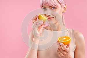 Portrait of smiling woman with pink hair holding half of ripe orange and smelling fresh lemon, posing isolated over pink