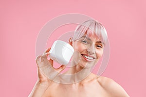 Portrait of smiling woman with pink hair holding white jar with moisturizing body lotion after shower isolated over pink