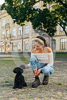 Portrait of a smiling woman sitting on the street and playing with a little black dog. Dog breed toy poodle. Vertical