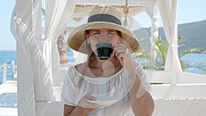 Portrait of smiling woman in straw hat drinking coffee in the sea beach cafe on a sunny windy day.
