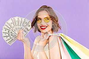 Portrait of a smiling woman in sunglasses holding shopping bags and money banknotes over violet