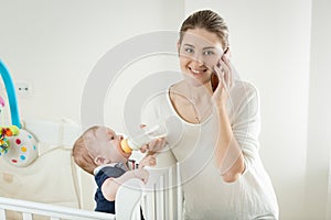 Portrait of smiling woman talking by phone while feeding her baby son