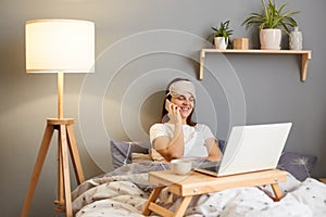 Portrait of smiling woman wearing sleep mask sitting with laptop in bed at home, watching movie and talking on mobile phone,