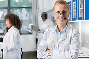 Portrait Of Smiling Woman In White Coat And Protective Eyeglasses In Modern Laboratory, Female Scientist Over Busy