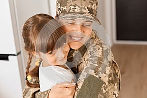 Portrait of smiling young adult soldier woman wearing camouflage uniform and cap posing with her daughter, hugging kid with