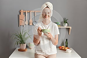 Portrait of smiling young adult woman wrapped white towel and dressed in T-shirt, doing cosmetology procedures and eating her