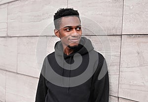 Portrait smiling young african man wearing black hoodie on city street over gray wall