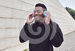 Portrait smiling young african man in wireless headphones enjoying listening to music wearing a black hoodie on a city street