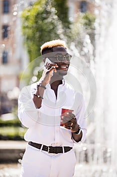 Portrait of smiling young afro american man talking on mobile phone and drinking phone while standing near the fountain in the cit