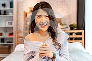 Portrait of smiling young Asian girl sitting on bed while making a video call in bedroom at home. Video conferencing