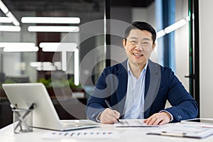 Portrait of a smiling young Asian man in a suit sitting in the office at a desk with a laptop and looking at the camera