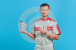 Portrait of smiling young asian mechanic pointing at plastic bottle of engine oil with finger over blue background