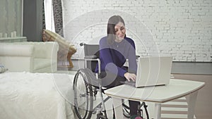 Portrait of a smiling young beautiful disabled woman in a wheelchair, working at home on a laptop, remote work on the