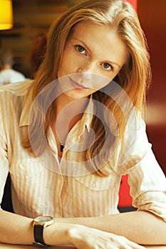 Portrait of smiling young beautiful red-haired student girl wearing trendy wrist-watch and casual shirt and posing in cafe.