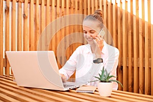 Portrait of smiling young beautiful woman freelancer works remotely on laptop in cafe, female talking on smart phone at work while
