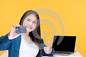 Portrait of smiling young beautiful woman holding a credit card in front of the laptop blank screen on yellow background. Shopping