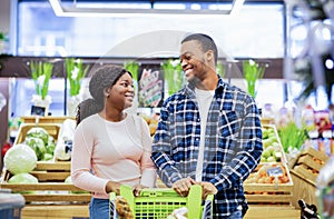 Portrait of smiling young black couple with shopping cart looking at each other in veggies section of grocery store