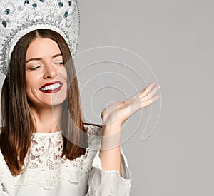 Portrait of a smiling young brunette woman in kokoshnik holding copy space on her palm isolated over grey background