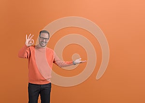 Portrait of smiling young businessman showing empty palm and OK sign on isolated orange background