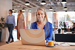 Portrait Of Smiling Young Businesswoman Sitting At Desk With Laptop In Modern Open Plan Office